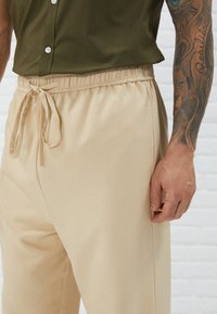 Beige elastiske bukser med snoreliv, glat tekstur og sidelommer, parret med en mørkegrøn kortærmet skjorte.