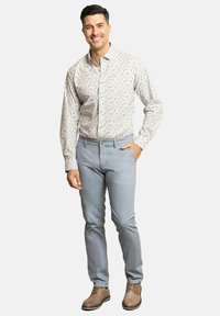 Camisa abotonada con una base beige clara y un patrón floral azul, combinada con pantalones gris claro y zapatos marrones de cordones. Pose de pie.
