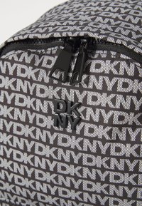 Fekete-fehér mintás hátizsák DKNY logóval, cipzárral és fényes fekete fém kiegészítőkkel. Anyaga tartósnak tűnik, texturált felülettel.