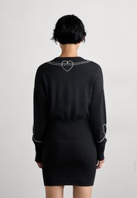 Fekete pulóver hosszú ujjakkal és bordázott passzéval. A háton és a mandzsettákon fehér szív alakú varrás található. Simább textúrájú.