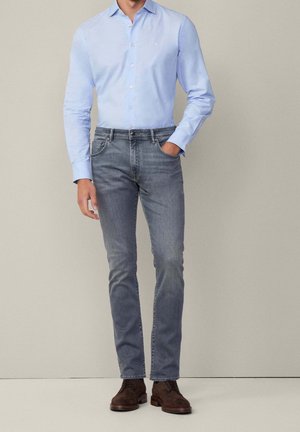 Homme portant une chemise bleu clair boutonnée, un jean gris slim-fit et des chaussures en cuir marron, debout avec une main dans la poche.