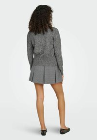 Pull en tricot gris à manches longues, associé à une jupe plissée à carreaux gris. Chaussures plates noires avec des clous blancs visibles.