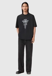 Czarny t-shirt z krótkim rękawem z misternym białym wzorem na środku, w połączeniu z luźnymi, ciemnymi dżinsami i czarnymi butami.