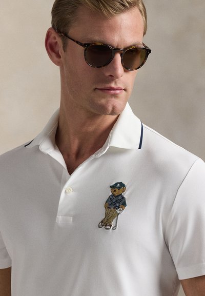 Λευκό πόλο μπλουζάκι με κέντημα αρκούδας σε γκολφ ντύσιμο στο αριστερό μέρος του στήθους. Η γιακάς έχει μπλε σκούρο περιθώριο. Το ύφασμα φαίνεται λείο και ελαφρύ.