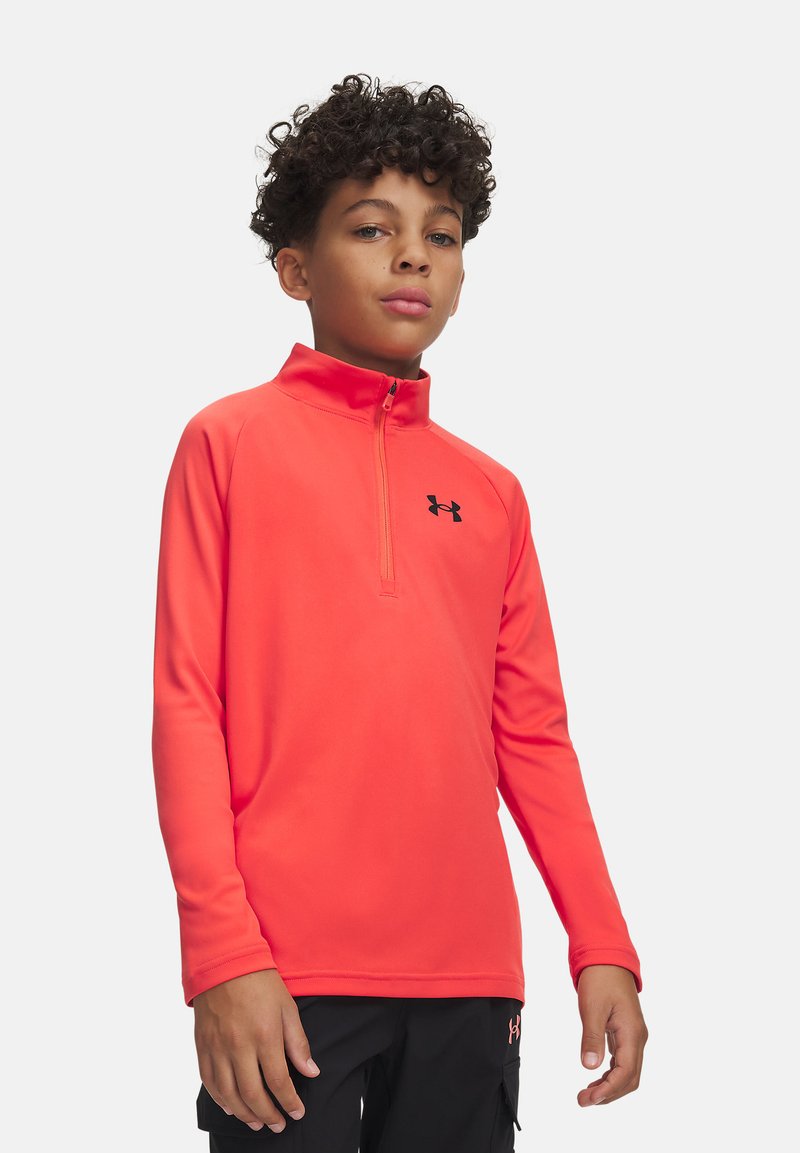 Garçon aux cheveux bouclés portant un haut corail vif à manches longues avec une fermeture éclair quart de zip et un pantalon noir, debout devant un fond clair.