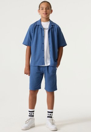 Garçon debout portant une chemise à manches courtes bleue avec boutons et un short assorti, un t-shirt blanc avec un motif graphique, des baskets blanches et des chaussettes noires et blanches.