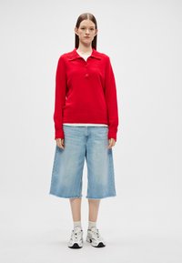 Červený pletený polo sveter s golierom a tromi gombíkmi, skombinovaný s svetlomodrými širokými džínsovými šortkami a bielymi teniskami.