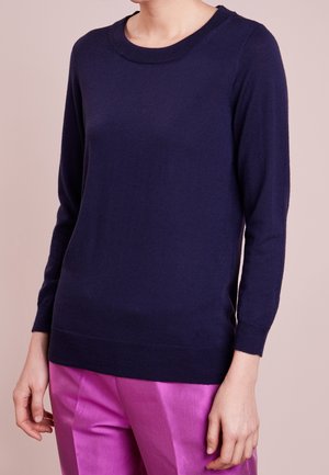 Slim fit donkerblauwe trui met lange mouwen gecombineerd met felroze satijnen broek bij een staande persoon tegen een neutrale achtergrond.