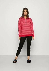 Kvinne står iført en rød mønstrete zip-up genser, svarte cropped bukser og svarte åpen-tå slippers mot en enkel bakgrunn.