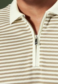 Polo rayé beige et blanc avec une fermeture à glissière sur le devant et un tricot doux et texturé. Caractéristiques : col contrastant et fermeture éclair métallique.