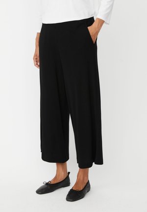 Persoon die zwarte brede cropped broek, zwarte ballerina's en een witte top met lange mouwen draagt, staand met één hand in de zak.