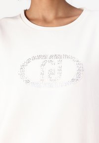 Bijela pamučna majica s širokim izrezom, koja sadrži iridescentni logotip ukrašen malim rhinestone-ima u ovalnom obliku.