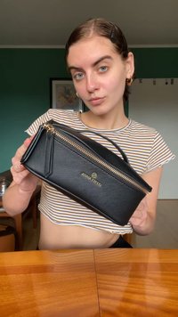 Giovane donna con i capelli corti e top corto a righe che tiene una borsa con zip nera di Anna Field con dettagli dorati in un ambiente domestico.