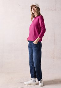 Fuchsia långärmad topp, avslappnad passform; kombinerad med blå jeans och vita sneakers. Modellen bär en ljusfärgad keps och står mot en neutral bakgrund.
