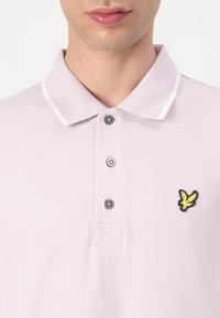 Homme portant un polo rose clair avec trois boutons et un logo d'oiseau jaune sur la poitrine gauche, lèvres et bas du visage visibles.