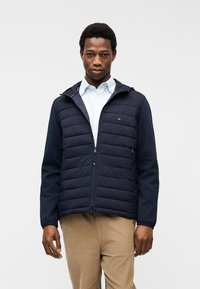 Άνδρας που φοράει ναυτικό μπλέ καπιτονέ μπουφάν πάνω από λευκό πουκάμισο και μπεζ παντελόνι, στέκεται μπροστά σε απλό ανοιχτό γκρι φόντο.