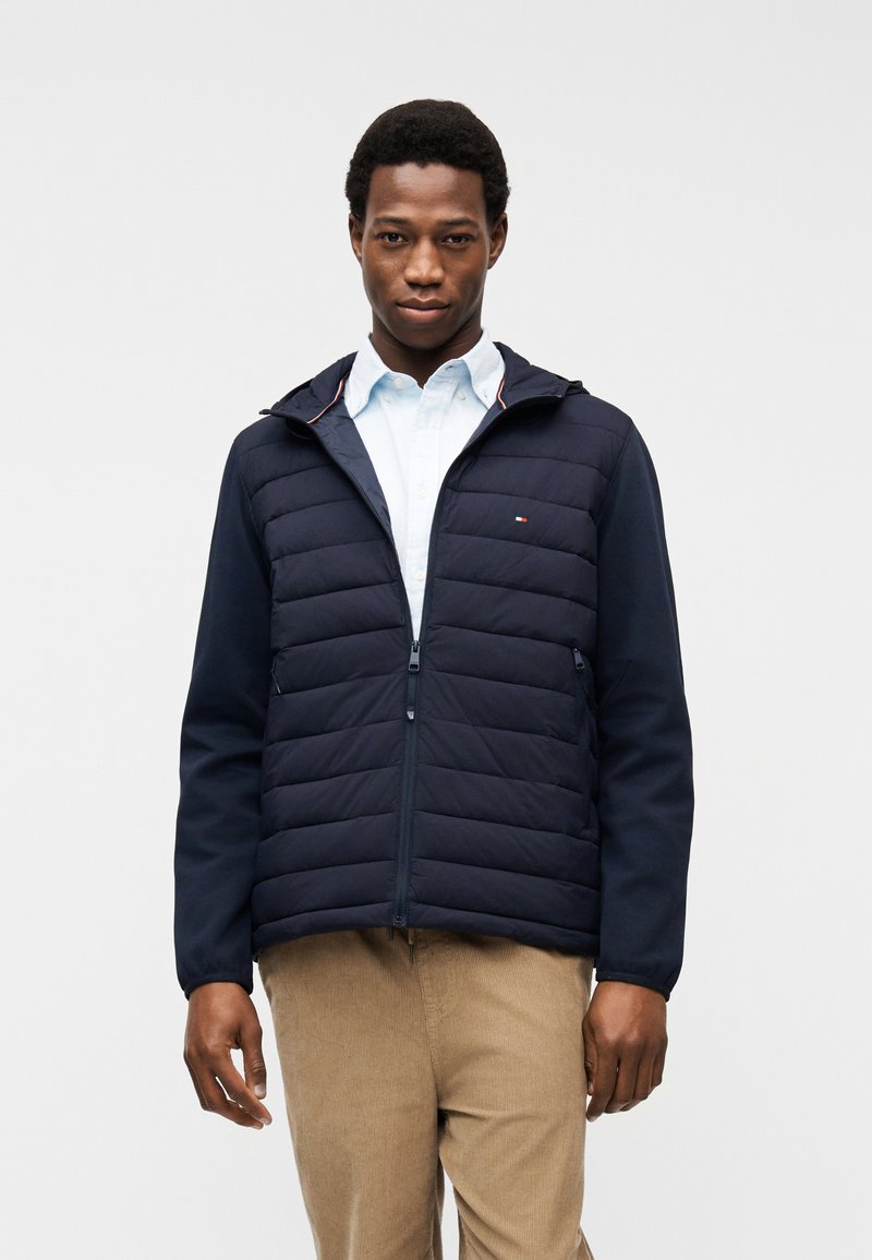 Άνδρας που φοράει ναυτικό μπλέ καπιτονέ μπουφάν πάνω από λευκό πουκάμισο και μπεζ παντελόνι, στέκεται μπροστά σε απλό ανοιχτό γκρι φόντο.