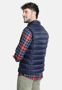 Chaleco de plumas azul marino con diseño acolchado, cuello alto y sin mangas, superpuesto a una camisa de cuadros en colores rojo, azul y verde.