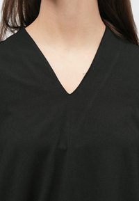 Melna v-veida kakla blūze no gludas auduma. Kakla izgriezums ir ass, un tekstūra šķiet mīksta ar minimāli redzamām šuvēm.