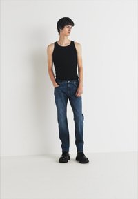 Musta ilmanhiha tank top yhdistettynä tummansinihin farkkuihin. Mallilla on päällään mustat paksupohjaiset kengät. Yksinkertainen, istuva malli valkoista taustaa vasten.