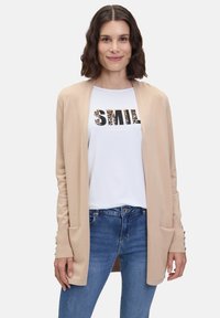 Beige cardigan met open voorkant, geribde manchetten en twee voorzakken, gedragen over een wit T-shirt met een "SMILE" graphic en blauwe spijkerbroek.