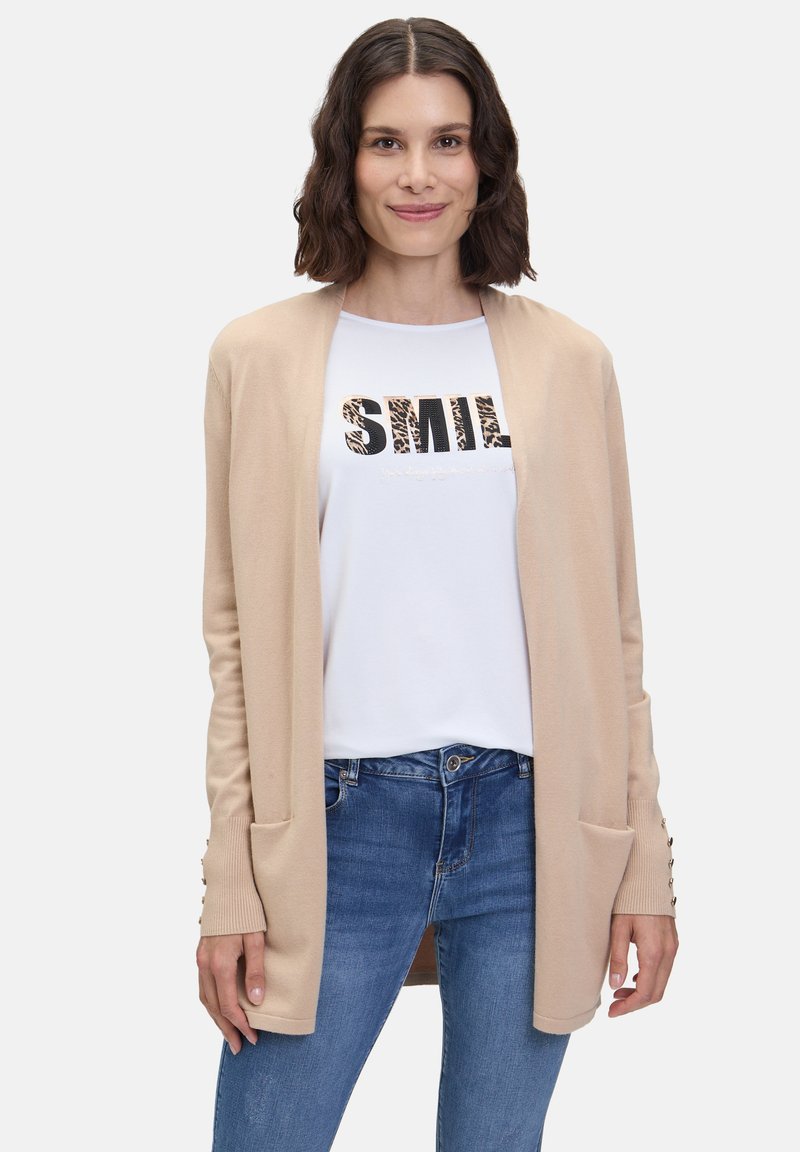 Beige cardigan met open voorkant, geribde manchetten en twee voorzakken, gedragen over een wit T-shirt met een "SMILE" graphic en blauwe spijkerbroek.
