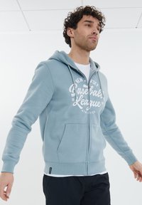 Helleblaue Zip-Hoodie aus weichem Stoff, mit einem Logo-Druck auf der Vorderseite, zwei Taschen und einer Kapuze mit Kordelzug. Modell in lässiger Pose dargestellt.