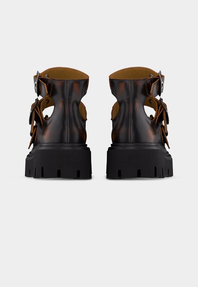 Χοντρά μποτάκια αστραγάλου από καφέ δέρμα με μαύρη σόλα, πλαϊνές αγκράφες και κοψίματα, σε γκρι φόντο, από πίσω.