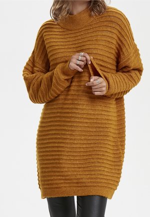 Femme portant une robe pull tricotée côtelée jaune moutarde, tenant le bord de la manche, associée à un pantalon noir et des bagues argentées.