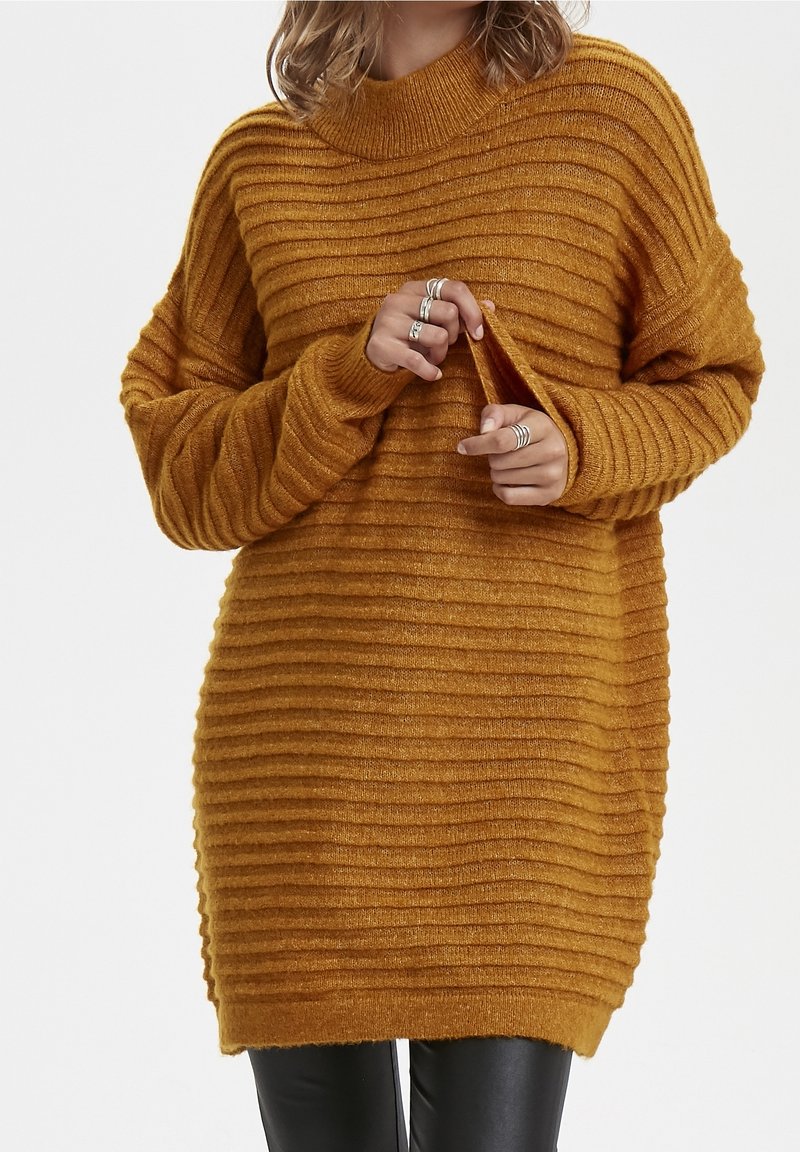 Femme portant une robe pull tricotée côtelée jaune moutarde, tenant le bord de la manche, associée à un pantalon noir et des bagues argentées.