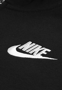 Fekete szövet, amelyen fehér hímzett Nike logó látható, jellegzetes ívelt pipa formával és szöveggel, puha tapintású és finom részletekkel.