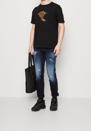 Sort bomulds t-shirt med gul grafisk design, blå slidte jeans og sorte sneakers. En kviltet sort tote taske holdes i den ene hånd.
