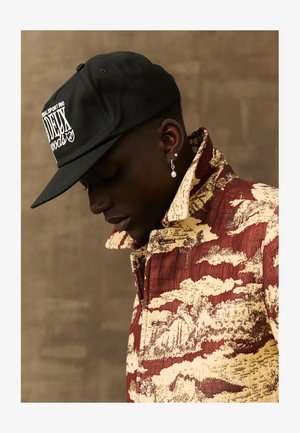 Camisa com padrão em tons de castanho-avermelhado e bege, com colarinho alto, combinada com um boné preto com logótipo branco. Um único brinco de pérola visível na orelha.