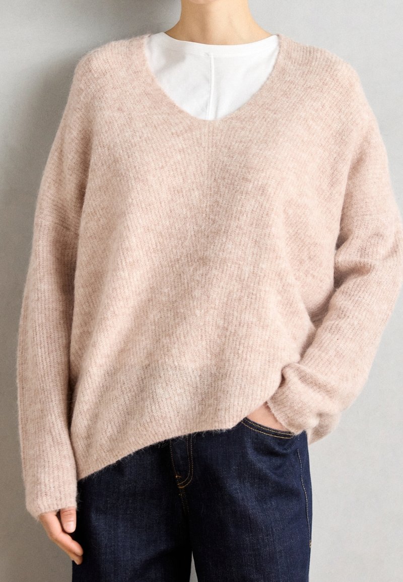 Pull doux rose pâle à texture duveteuse avec col en V porté sur une chemise blanche, assorti à un jean bleu foncé, main dans la poche, sur fond uni.