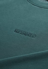 Sudadera azul verdosa con cuello acanalado, con un logo "SUPERDRY" en relieve. Hecha de tela suave, con una textura lisa y costuras resistentes.