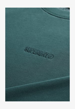 Bluza w kolorze morskim z prążkowanym kołnierzem, ozdobiona wytłoczonym logo „SUPERDRY”. Wykonana z miękkiego materiału, o gładkiej fakturze i solidnym szyciu.