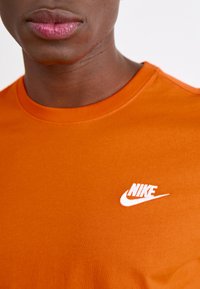 Oranžs Nike T-krekls ar apaļu kaklu, īsām piedurknēm un baltu Nike logo uz krūtīm. Gluda auduma tekstūra.