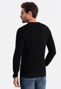 Schwarzer gestrickter Pullover mit einem strukturierten Muster, rundem Ausschnitt sowie gerippten Bündchen und Saum, kombiniert mit blauen Jeans.