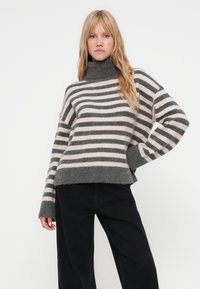 Stripet genser med høy hals i grått og blush, med løs passform, vide ermer og myk tekstur. Kombinert med svarte bukser.