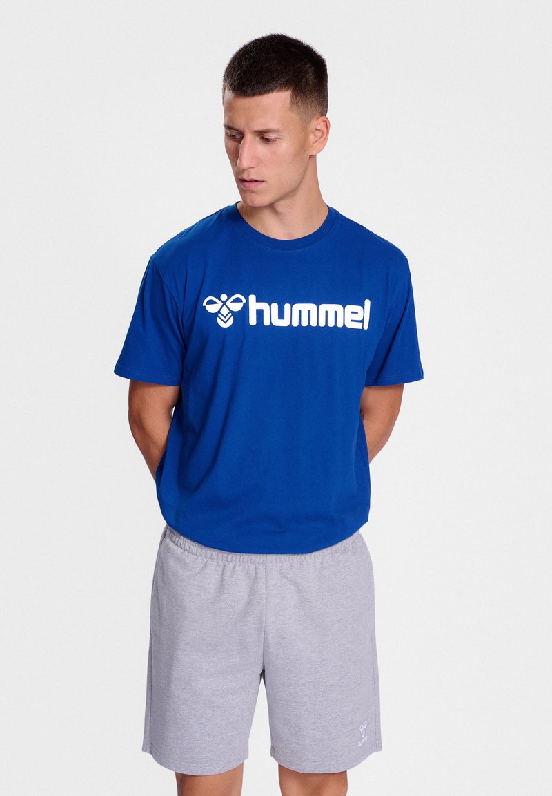 Blå bomullströja med vitt "hummel"-logga, parat med grå shorts. Avslappnad design, korta ärmar och en avslappnad passform.
