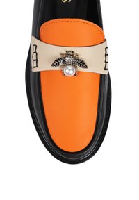 Skórzane mokasyny z czarną podstawą, jasnopomarańczową cholewką oraz kremowym paskiem ozdobionym pszczółką z perłą w centrum.