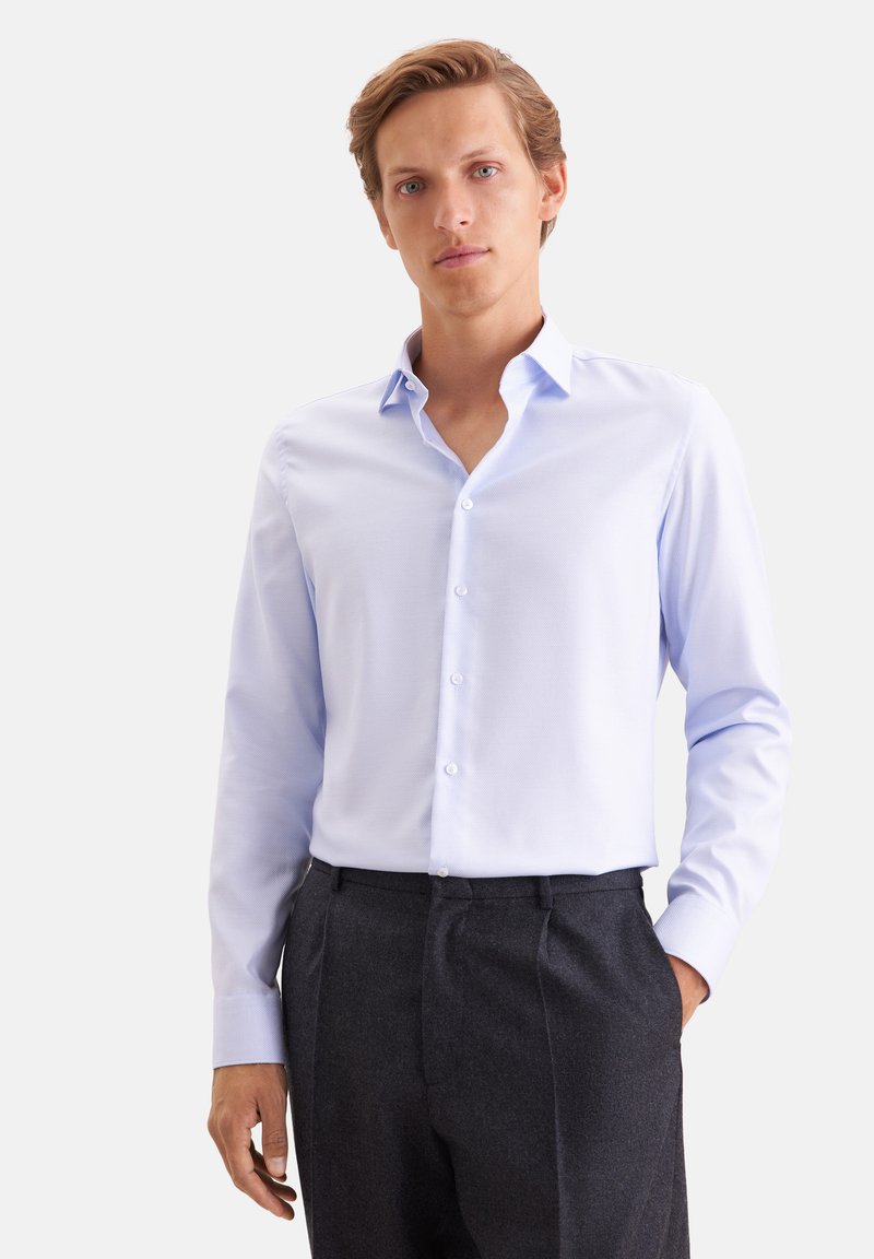 Jeune homme aux cheveux châtain clair portant une chemise bleu clair à manches longues boutonnée et un pantalon gris foncé, debout avec une main dans la poche.
