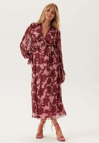 Kvinna i en långärmad, rosa och vinröd blommig midiklänning med volanger, ihopparad med rosa snörda högklackade sandaler, stående och leende.