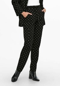 Woman wearing black tapered pants with white polka dots, matching black polka dot blazer, white top, and black heeled ankle boots, hands in pockets.
