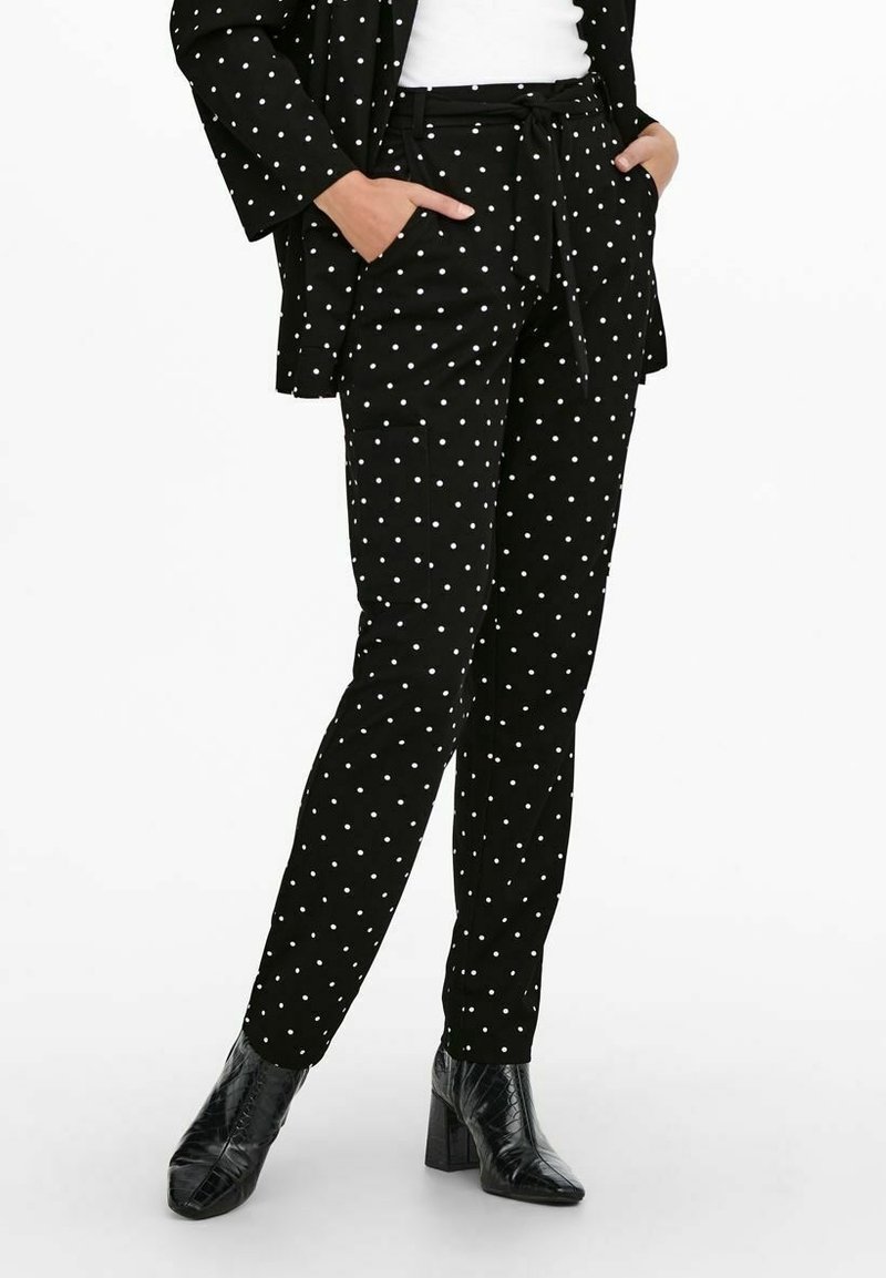 Woman wearing black tapered pants with white polka dots, matching black polka dot blazer, white top, and black heeled ankle boots, hands in pockets.