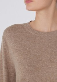 Gros plan d'une personne portant un pull-col rond marron clair, montrant le cou, le menton et une partie du visage tourné sur le côté.