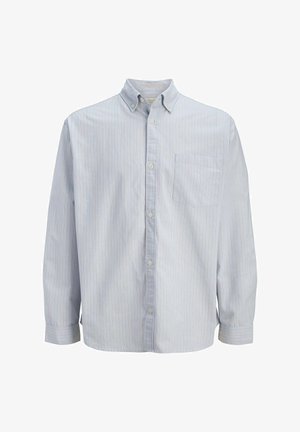 Garçon debout, portant une casquette marron, un T-shirt rouge à motif, une chemise bleu clair ouverte à boutons, un jean délavé à jambes larges, et des baskets blanches.