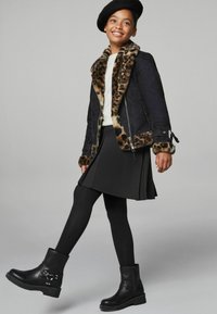 Sort jakke med leopardprint faux fur krave og manchetter, cremefarvet sweater, sort plisseret nederdel og sorte ankelstøvler med nitter.
