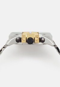 Orologio argento con quadrante rettangolare, dettagli dorati sulla corona, pulsanti neri e cinturino traforato in argento. Finitura in metallo lucido.