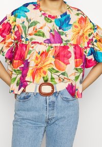 Blomstret bluse med flæseærmer, prydet med flerfarvede liljer og blade på en lys baggrund, sat sammen med lyseblå jeans og et beige bælte.